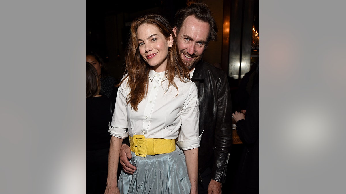 Michelle Monaghan in a white blouse with a yellow belt and blue skirt held by her husband in a leather jacket